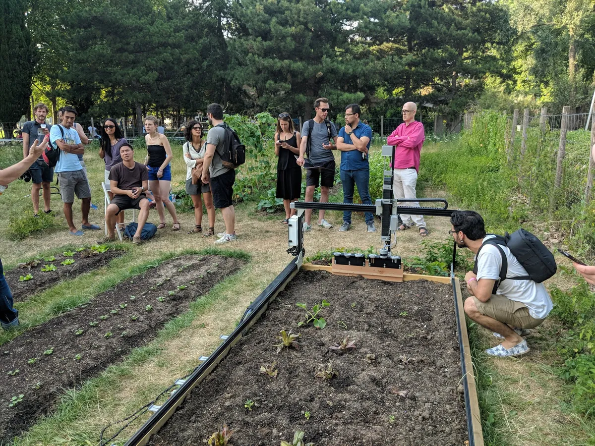 A group of neighbors gathered around community garden beds while a FarmBot tends a row of seedlings